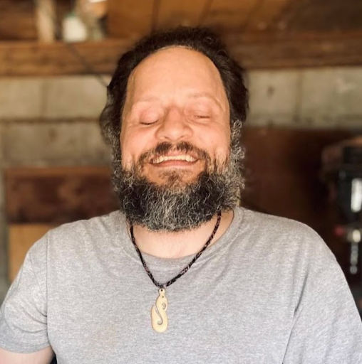 A man with a beard and mustache smiles with his eyes closed. He is wearing a light gray t-shirt and a necklace with a pendant. The background appears to be an indoor space with wooden elements.